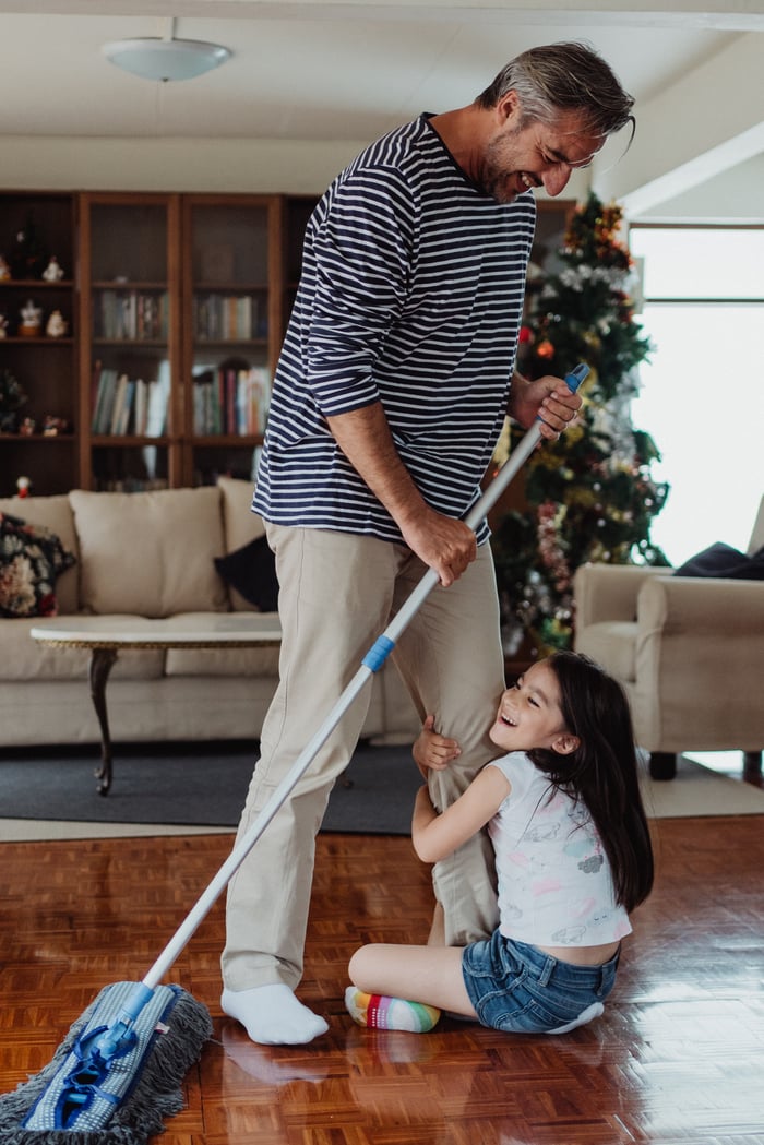 portfolio-02 A father and his daughter having fun together while mopping the living room floor, near a decorated Christmas tree.