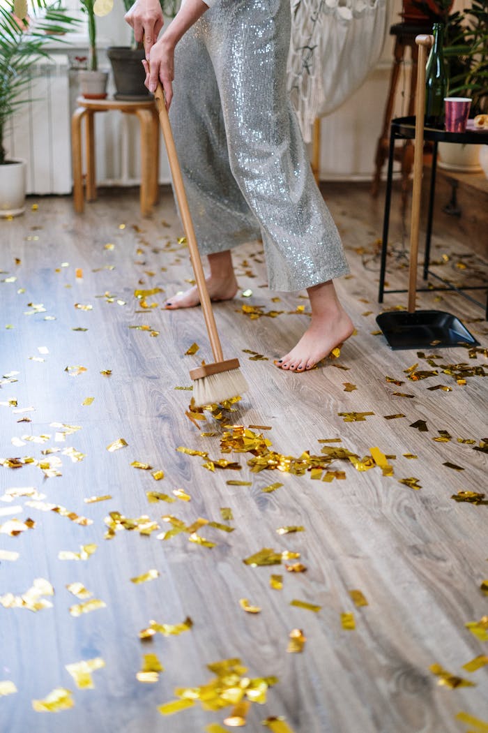 portfolio-05 Person sweeping golden confetti off wooden floor after a party, barefoot indoors.